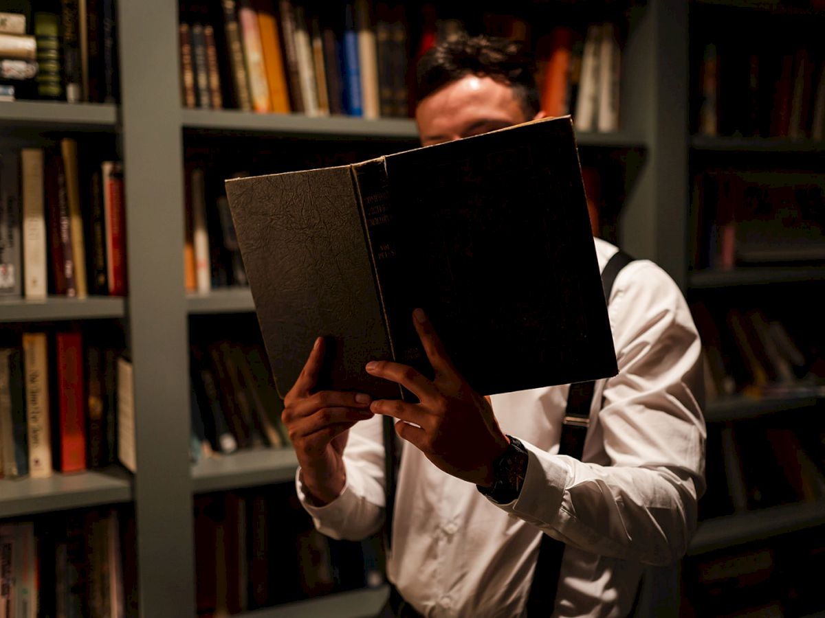 A person in a white shirt holds an open book in front of their face, standing in a dimly-lit library with shelves of books.