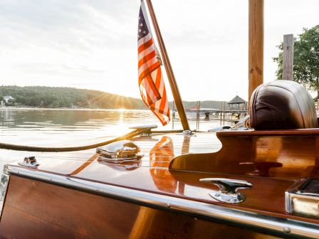 A close-up of a wooden boat with an American flag at sunset, docked by a tranquil lake with trees and a pier in the background.