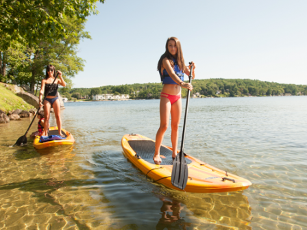 Two women are paddleboarding on a calm body of water near a shore with greenery.