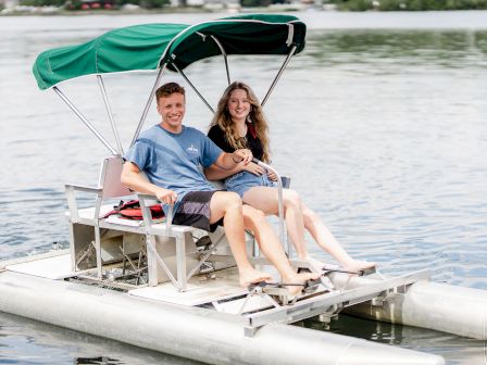 A smiling couple is enjoying a ride on a pedal boat with a green canopy, floating on calm waters with a scenic background.