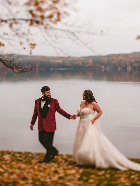 A couple in wedding attire walks hand-in-hand near a lake, surrounded by autumn leaves, with hills and colorful trees visible in the background.