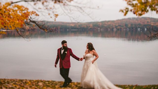 A couple in wedding attire walks hand-in-hand near a lake, surrounded by autumn leaves, with hills and colorful trees visible in the background.