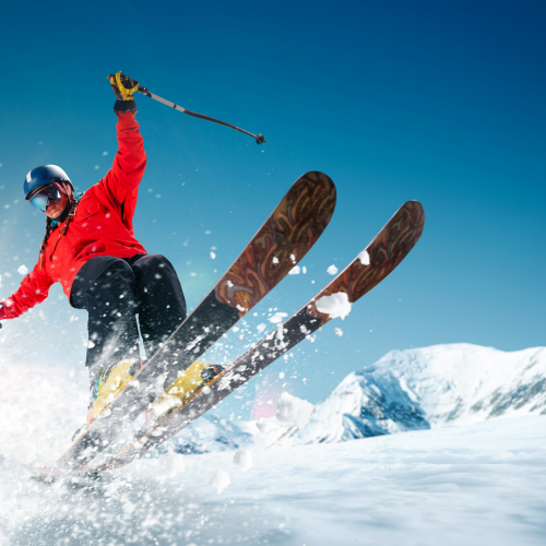 A skier in a red jacket and black pants is jumping in the air on snowy slopes with mountains in the background and a clear blue sky.