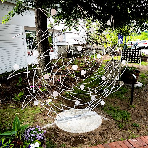 The image shows a metal sculpture with abstract designs, situated outdoors near a brick pathway and surrounded by small plants and flowers.