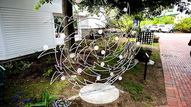 The image shows a metal sculpture with abstract designs, situated outdoors near a brick pathway and surrounded by small plants and flowers.