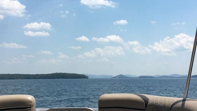 The image shows a serene lake view with scattered clouds in the sky, seen from the seating area of a boat, with islands in the distance.