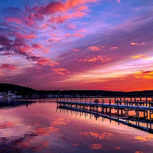 A serene lake with a wooden dock extending into the water, under a vibrant sunset sky with hues of pink, purple, and orange, ending the sentence.