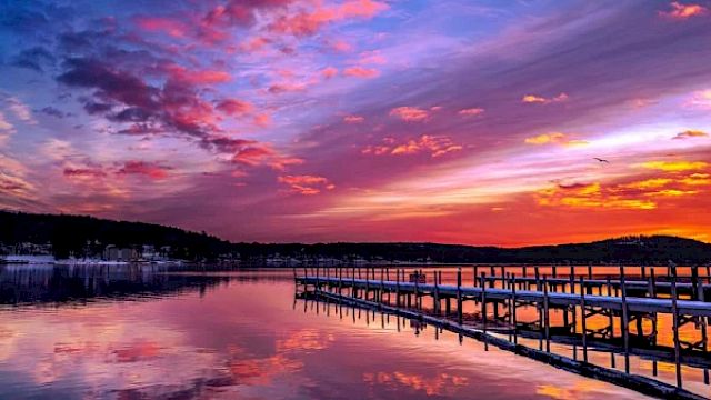 A serene lake with a wooden dock extending into the water, under a vibrant sunset sky with hues of pink, purple, and orange, ending the sentence.