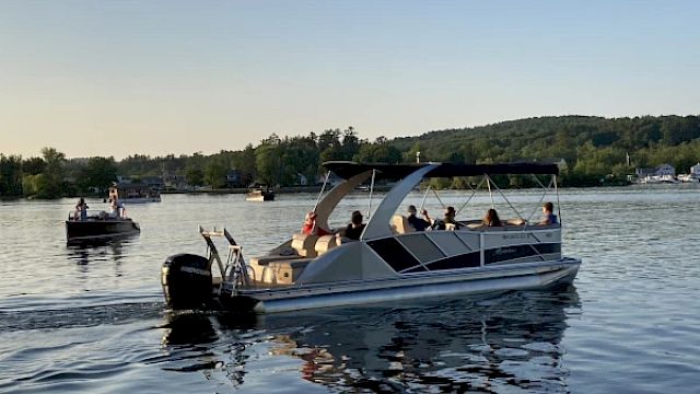 A pontoon boat filled with people is cruising on a calm lake with a distant shoreline and other boats in the background during sunset.