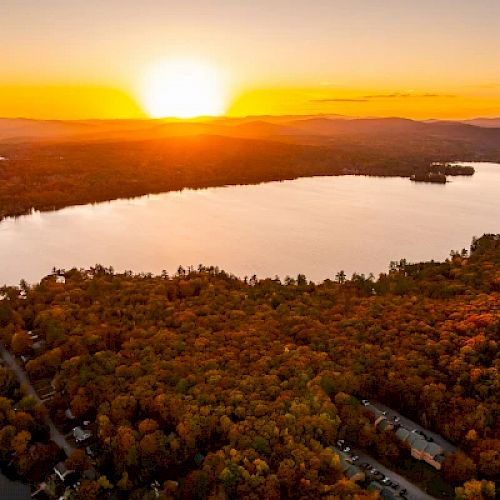 A stunning sunset over a large lake, surrounded by dense forests with autumn foliage and hills in the background, creating a serene landscape.