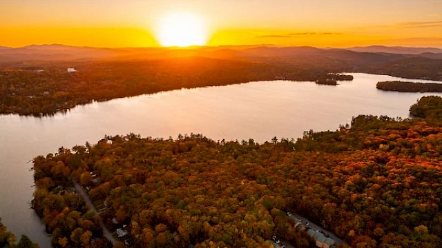 A stunning sunset over a large lake, surrounded by dense forests with autumn foliage and hills in the background, creating a serene landscape.