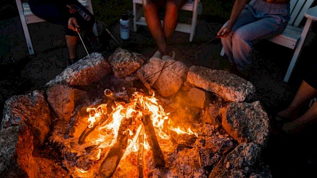 A group of people sitting around a campfire, roasting marshmallows with sticks, surrounded by rocks and seated in chairs.