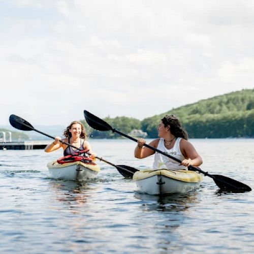 Two people kayaking on a lake with lush green hills in the background, both paddling with black oars and enjoying the water.