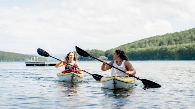 Two people kayaking on a lake with lush green hills in the background, both paddling with black oars and enjoying the water.