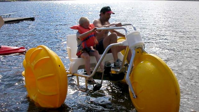 A man and a child are on a yellow water tricycle, pedaling on a lake with life vests on.