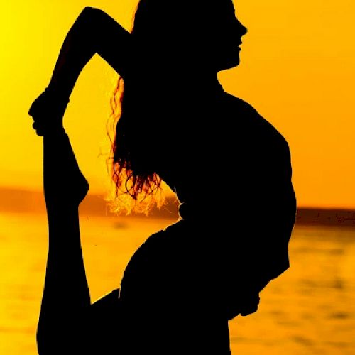 A silhouetted person is performing a yoga pose by a body of water during sunset, with their leg held up behind them in a graceful stretch.