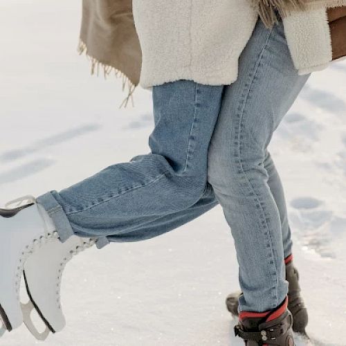 Two people are standing on ice; one person is wearing ice skates while the other person is wearing boots, both are dressed warmly.