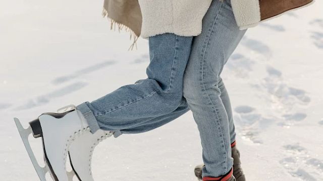 Two people are standing on ice; one person is wearing ice skates while the other person is wearing boots, both are dressed warmly.