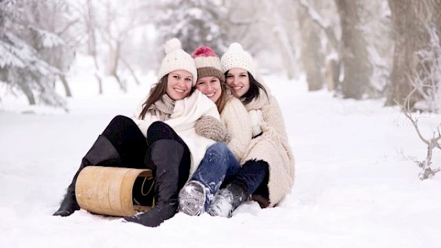 Three people in winter clothing are sitting together on the snow with a wooden sled nearby, smiling and enjoying the snowy environment.