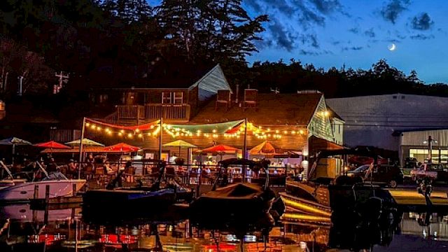 A lively waterfront scene at dusk with string lights, umbrellas, boats, and a crescent moon in the sky, reflecting on the water.