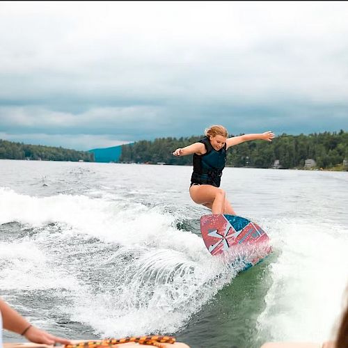 A person is wakeboarding on a lake, performing a trick on a wave behind a boat, with two people watching in the foreground.