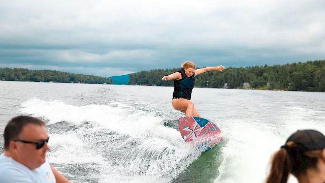 A person is wakeboarding on a lake, performing a trick on a wave behind a boat, with two people watching in the foreground.