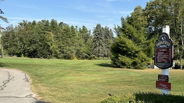 A paved path next to a lawn, trees, and a sign under a clear blue sky.