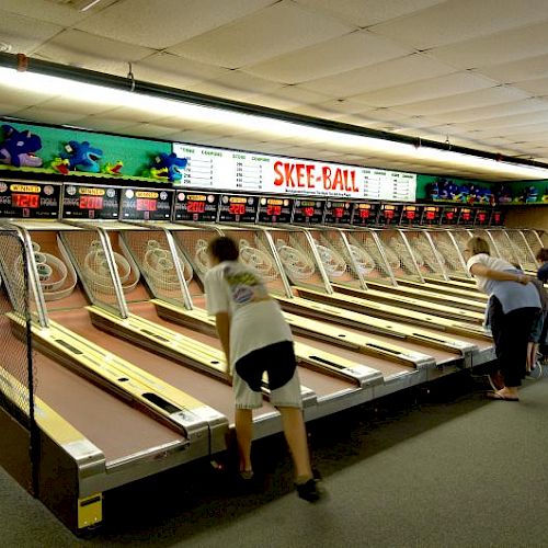 People playing skee-ball in an arcade lined with multiple alleys and a colorful sign above the lanes.