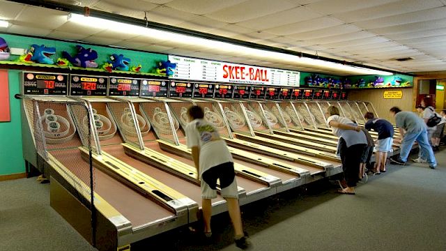 People playing skee-ball in an arcade lined with multiple alleys and a colorful sign above the lanes.