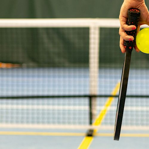 A hand holding a pickleball paddle and a yellow pickleball on an indoor court with a net in the background.