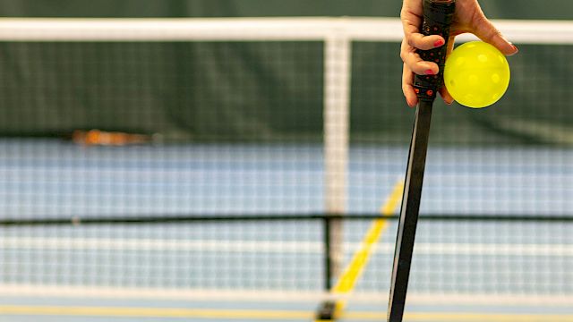 A hand holding a pickleball paddle and a yellow pickleball on an indoor court with a net in the background.