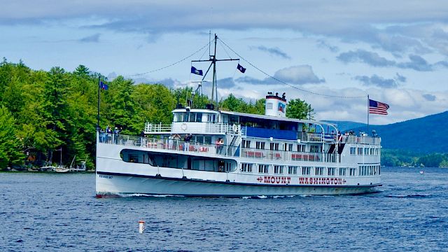 A white boat named Mount Washington cruises on a lake, with a U.S. flag atop. Tree-lined shore and hills are visible in the background.