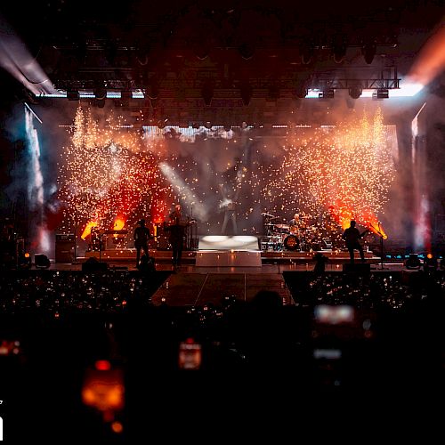 A concert stage with vibrant lighting and sparkling pyrotechnics at BankNH Pavilion, viewed by an audience holding up phones.