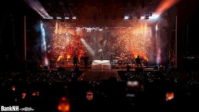 A concert stage with vibrant lighting and sparkling pyrotechnics at BankNH Pavilion, viewed by an audience holding up phones.