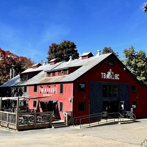 A red barn-like brewery building surrounded by trees, with outdoor seating and clear blue skies above.