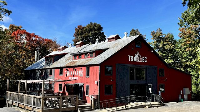 A red barn-like brewery building surrounded by trees, with outdoor seating and clear blue skies above.