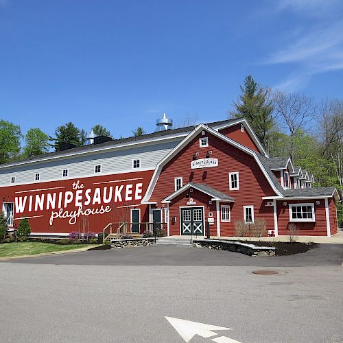 A red building labeled "WinniPesaukee Playhouse" set in a wooded area under a clear blue sky, with a road and parking area in front.