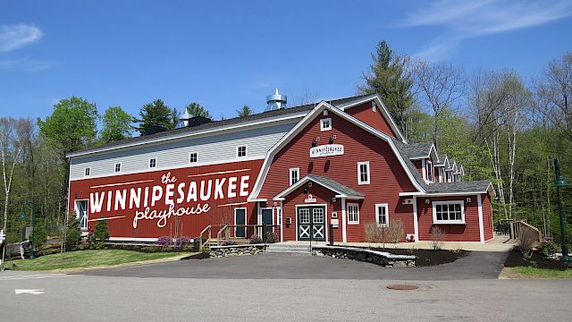 A red building labeled "WinniPesaukee Playhouse" set in a wooded area under a clear blue sky, with a road and parking area in front.