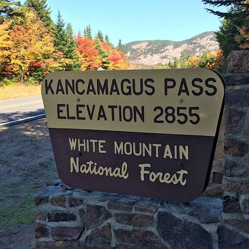 A sign for Kancamagus Pass, elevation 2855, in White Mountain National Forest with a road and colorful autumn trees in the background.