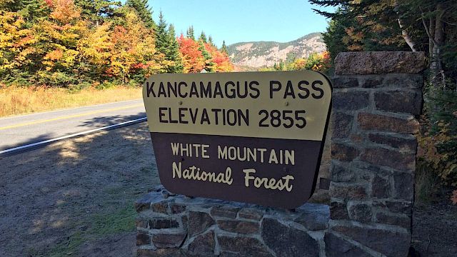 A sign for Kancamagus Pass, elevation 2855, in White Mountain National Forest with a road and colorful autumn trees in the background.