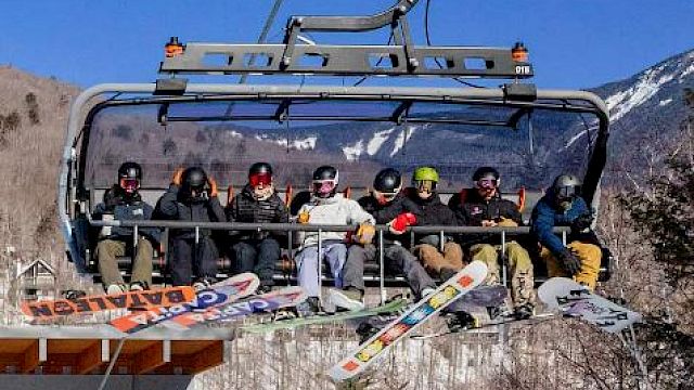 A group of people on a chairlift with snowboards in a mountainous, snowy area, enjoying a sunny day outdoors.