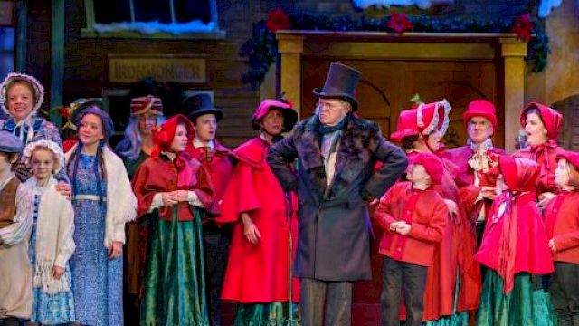 A group of people in period costumes on stage, possibly from a holiday-themed play, with festive decorations in the background.