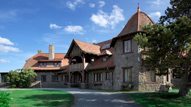 A large stone house with a rustic design, featuring steep roofs and a tower, surrounded by greenery and a clear blue sky.