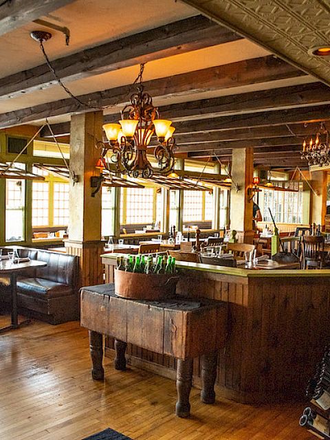 The image shows a cozy restaurant interior with wooden beams, chandeliers, and cushioned booths. Wine bottles are displayed on a rack.