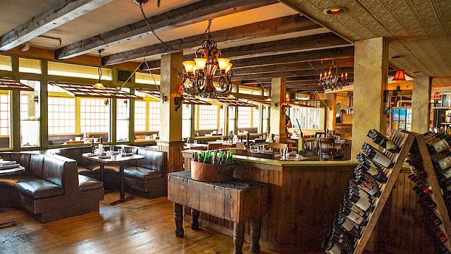 The image shows a cozy restaurant interior with wooden beams, chandeliers, and cushioned booths. Wine bottles are displayed on a rack.