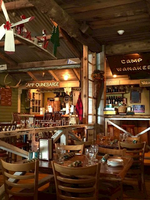 The image shows a cozy, rustic restaurant interior with wooden furniture, vintage decor, and signs like "Camp Wanakee" and "Watch Yer Step."