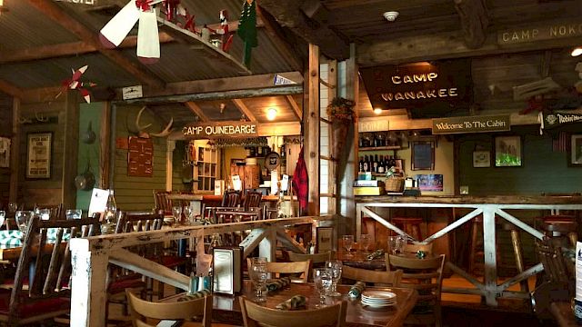 The image shows a cozy, rustic restaurant interior with wooden furniture, vintage decor, and signs like "Camp Wanakee" and "Watch Yer Step."