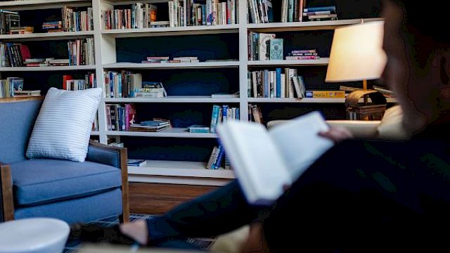 A person reads a book in a cozy room with full bookshelves, an armchair, and a lit lamp, creating a peaceful atmosphere.