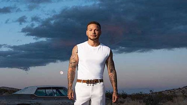 A person in a white outfit walks in a desert landscape at dusk, with a classic car and cloudy sky in the background.