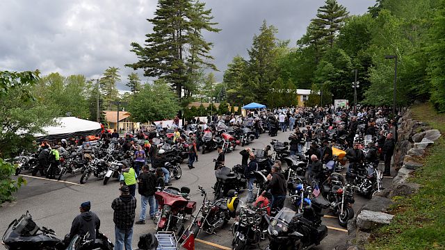 A large gathering of people and motorcycles in a parking area surrounded by trees under a cloudy sky.